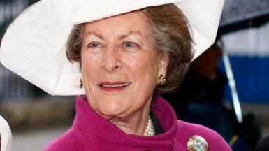 Lady Pamela Hicks in a pink coat with a gold brooch on the shoulder, and a large white hat. 
