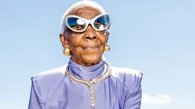 Margret Chola in a shiny lilac top wearing cat-eye mirrored sunglasses, a bejewelled gold necklace  in the shape of a snake and big gold ball earrings