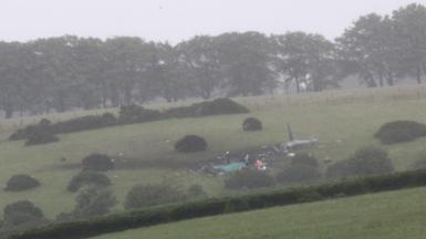 Scene of a helicopter crash in a field. Debris from the grey military helicopter is strewn across the grass. Wreckage can be seen across parts of the field. Tarpaulin is on top of the wreck. 