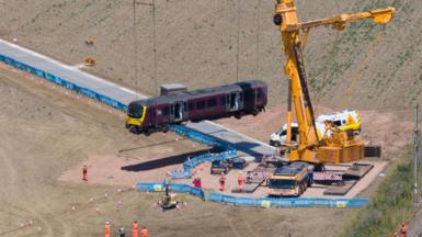A giant yellow crane in a field next to railway line. It is lifting a train carriage. A temporary track has been built to get the crane into position in the field. There are about a dozen people in orange overalls.