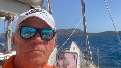 Middle-aged man on a sailing boat faces the camera wearing black sunglasses with reflective lenses, an orange top and white cap 