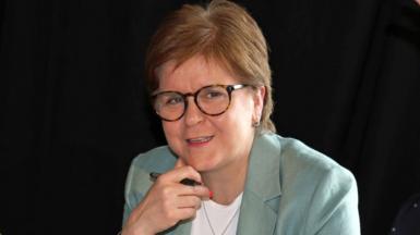 Nicola Sturgeon sitting down, wearing a pale green suit over a white top. She has her hand underneath her chin and is holding a pen.