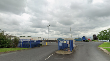 The main entrance of Wetherby-based Moores Furniture Group. It shows buildings in the distance and in the foreground blue gates and a security hut with a number of large trucks bearing the Moores logo parked behind the main gate