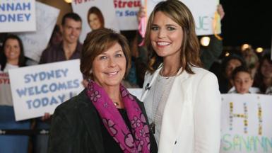 Savannah, all in white, and Nancy Guthrie in a black suit jacket and pink patterned scarf, smile as they stand shoulder to shoulder. People behind them hold up signs. 