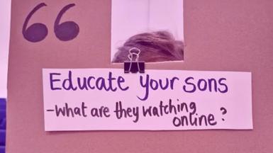 A cardboard sign is held up to the camera, in marker pen it says 'Educate your sons - what are they watching online?' 