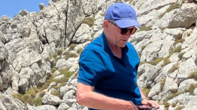 Michael Mosley on a beach, he is wearing a blue cap, sunglasses and a blue Polo shirt