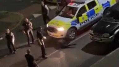 A number of police officers in black uniforms next to a police car. The cow is stuck underneath the police car. 