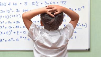 A child in a white school shirt faces away from the camera with his hands clasped to the back of his head. He is looking at a school whiteboard littered with maths equations in blue ink, set against a green wall. 