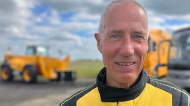 A man looks towards the camera wearing a yellow and black driving suit with the "JCB" logo on the front.