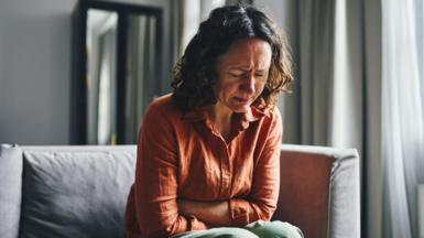Stock image of woman with curly brown shoulder length hair sitting on a grey softa. Her arms are folded over her abdomen and she appears to be grimacing and upset in pain. She wears a terracotta shirt and green trousers. There is light coming in through a nearby window. 