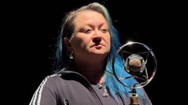 A woman with long grey and blue hair standing at a stage microphone. She's wearing a dark grey tracksuit top