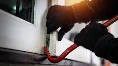 Gloved hands holding a crowbar being used to prise open a window