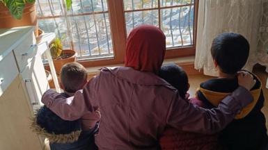 Niluper in a red headscarf and a mauve jacket with her arms around her three children, all of them in winter jackets - their backs are to the camera as they look out sit on the floor and look out the window.   