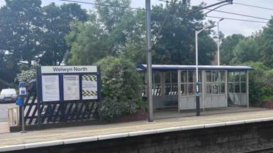 Welwyn North railway station, with a shelter to the left, railway tracks in front, a platform and trees behind the shelter. A sign is to the left. 