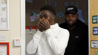 Bukayo Saka in a white zip-up shirt stands nervously, with hands covering his mouth, in a classroom setting.