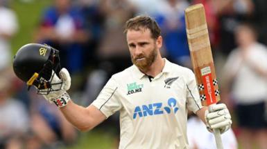 Kane Williamson, wearing New Zealand whites, holds his black helmet in his right hand and his cricket bat in his left to acknowledge a century