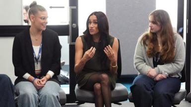 The Duchess of Sussex speaks to young people at a university - they are sit in a semi circle and Meghan is gesturing as she speaks.