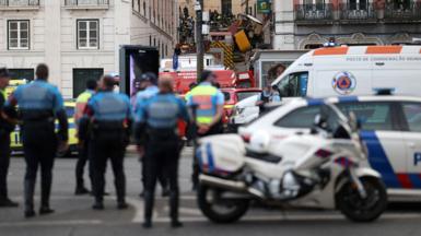 Police officers stand by near the scene of the crash with the overturned funicular seen crashed in the street above