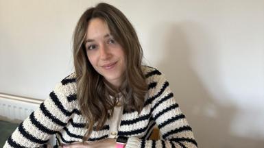 Abbi, a young woman with dark blonde hair. She is wearing a black and white striped cardigan and is sitting at her kitchen table