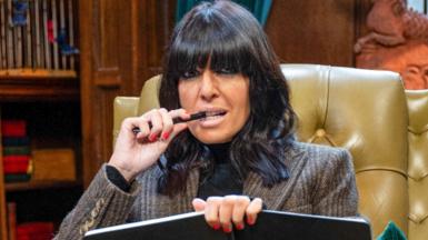 Claudia Winkleman on The Traitors, holding a notebook and pen