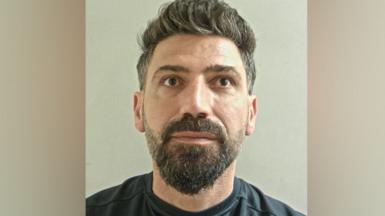 Amanj Hasan Zada's mugshot. He has a greying brown beard and wears a grey/blue t-shirt.