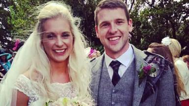 Anneka Johnstone and Alan Johnstone on their wedding day. Anneka has long blonde hair blowing in the wind and is wearing a white off-the-shoulder wedding dress, whilst Alan has short dark hair and is wearing a kilt jacket with a tartan shawl.