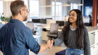 A young woman wearing a grey jumper over a shirt and blue jeans shaking hands with a man wearing a blue shirt and glasses in an office 