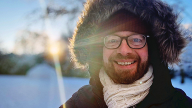 BBC reporter Ken Banks in snow, smiling at camera, wearing a thick hooded jacket with a fur trim round the hood and a scarf around his neck. The sun is shining in the background.