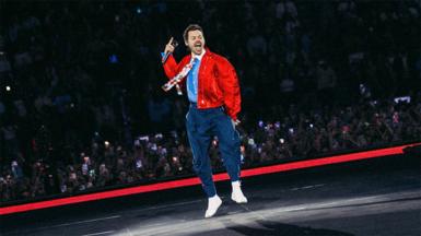 Harry Styles is wearing navy trousers with a belt and a light blue shirt with a white collar and flower patterned tie.He has a red bomber jacket over the top. He is jumping on the stage, you can see fans watching from the crowd behind him.