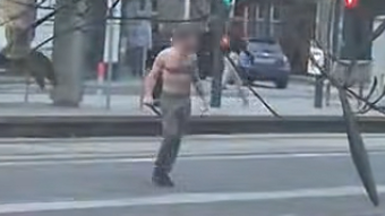 A still from a video showing a bare-chested man wearing dark trousers standing in the middle of a street. He is carrying a weapon in his hand. The still is blurry and the man's face has been delibrately blurred.
