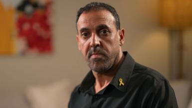 Eli Sharabi, wearing a black shirt, looks at the camera with little expression as he sits in front of a sofa, wall painting and lamp, all blurred in the background