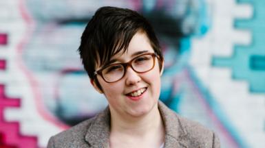 Lyra McKee stands with her arms folded in front of a brightly painted wall.
She is a young woman with short black hair, with a fringe over her forehead coming to rest on her glasses. She is smiling
She is wearing a brown tweed jacket over a white a navy striped top.