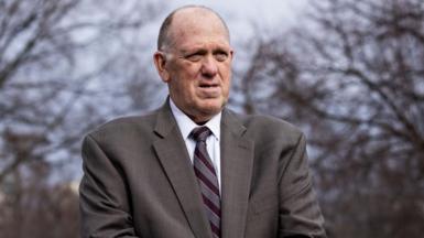 Tom Homan stands outside in front of leafless trees wearing a grey suit.