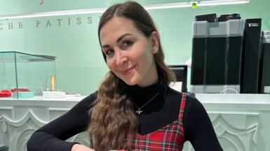 A woman with long brown hair, wearing a black turtle neck top and plaid dress smiling. 