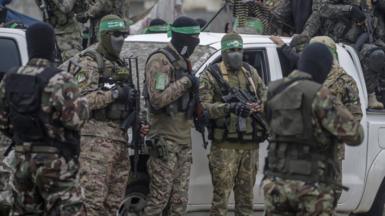 Armed members of Hamas stand in camouflage uniforms in front of a white car. Their faces are covered by balaclavas.