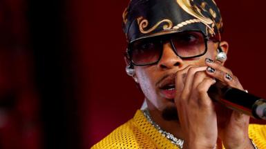 D4vd sings into a microphone. He wears a yellow sports jersey, lots of diamond jewelry, a silk scarf tied around his head and sunglasses
