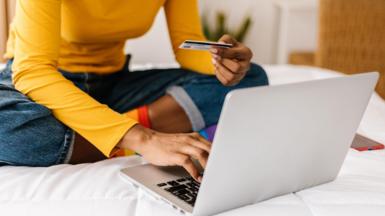 Woman in yellow top sits crossed legged on a bed while holding a credit card and typing on her laptop