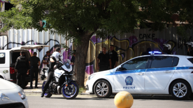 A police care and police officer astride a motorbike in a city street. There is a corrugated fence in the background covered in graffiti.