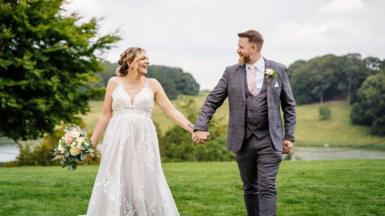 A newlywed couple hold hands and smile at each other against a backdrop of hills and trees. The groom is wearing a grey suit and the bride, who is holding some flowers in her other hand, is wearing a traditional white dress.