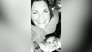 A woman smiles up at the camera. She is taking a selfie with a young boy who is also smiling at the camera. The photo is in black and white.