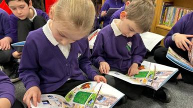 A young girl and a boy sit in their classroom on the floor both reading a book. They are in uniforms of a purple jumper or cardigan, white shirt and black trousers