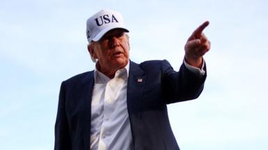 Trump wears a white USA cap and a dark suit, while pointing at something off-camera