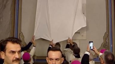 The Pope and church staff tug at white fabric covering a plaque inside a church