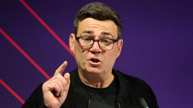 Andy Burnham, dressed all in black and wearing glasses, raises a finger to make a point during a speech.