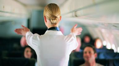 An air stewardess facing away from the camera shows passengers on a jet the emergency exist during a safety briefing. She is wearing a white shirt and her blonde hair is in a tight bun