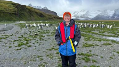 Aoife McKenna in Antarctica. She is wearing a red winter hat, glasses, and blue coat, with penguins and snowy hills behind her