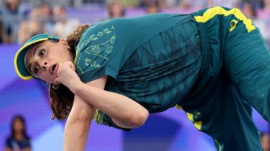 Breakdancer Raygun holds herself up on one arm, while the other crosses in front of it to allow her to hold her chin. She wears the green and gold of the Australian Olympic team. In the background are seated spectators.