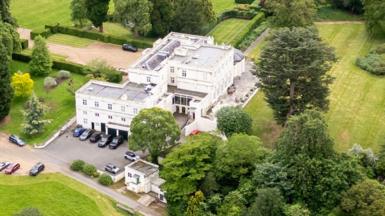 An aerial photograph of Royal Lodge, Prince Andrew's residence in Windsor, shows a large white building with green fields and trees surrounding it and a variety of cars parked outside it. 
