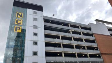 A multi-story car park with brick and glass frontage with the letters N C P on the glass section against the backdrop of a cloudy sky.