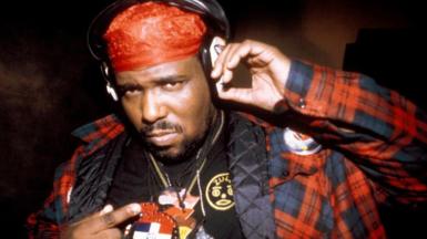 Afrika Bambataa poses with headphones and a red bandana as he points to emblems on his necklaces and shirt 